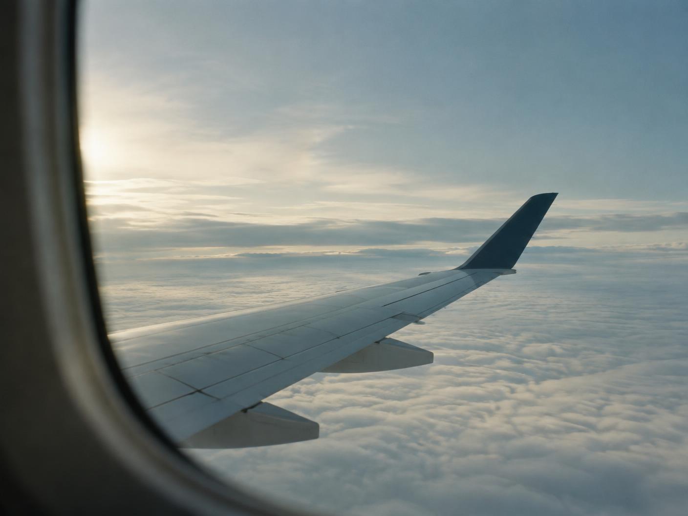 The wing of an airliner seen through a cabin window, above a flat layer of cloud in soft daylight.